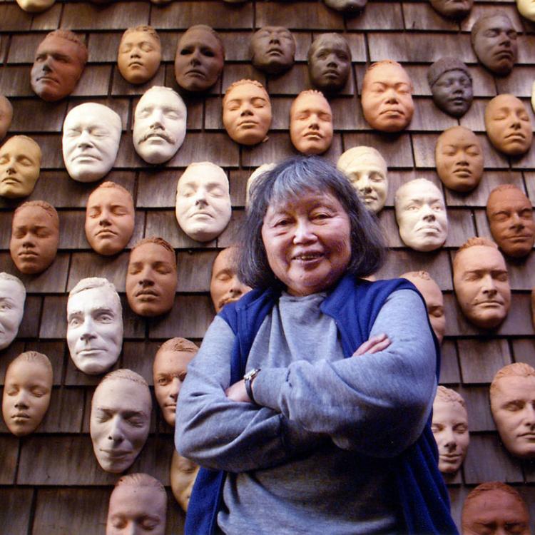 An image of artist Ruth Asawa infront of her Wall of Masks