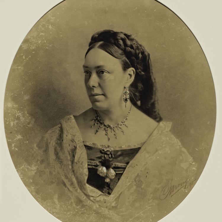 Short Alt Text A sepia-toned oval studio portrait of a woman looking to the side, wearing a formal off-the-shoulder dress, fine jewelry, and an elaborate braided hairstyle.
