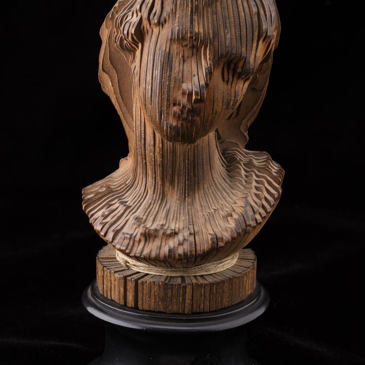 A wooden portrait bust sculpture set against a plain black background, mounted on a round black pedestal. The sculpture is constructed from numerous thin, vertical wooden slats arranged radially around a central axis, bound together near the top and base with thin light-colored cord. The carved edges of the slats collectively form the detailed contours of a person's head, face, and shoulder area.