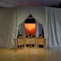 Panoramic view of a dimly lit installation featuring a large canvas tent with three openings. The polished wooden floor reflects the space. Each opening contains a screen displaying a moody, ominous landscape. In the center opening, a semicircular bar with three stools holds three VR headsets.