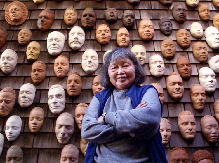 An image of artist Ruth Asawa infront of her Wall of Masks