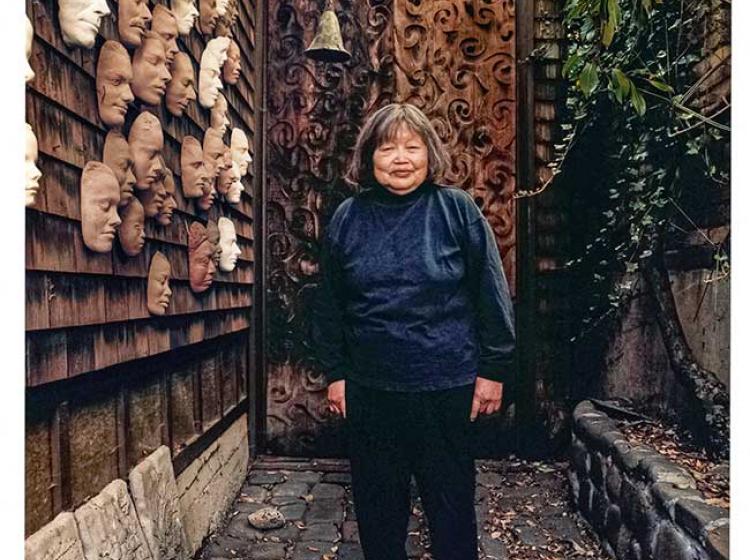 An image of Ruth Asawa standing outside her house next to her wall of masks.