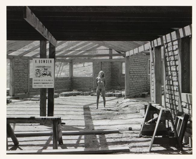A black and white photograph depicting a construction site, with a poster in the foreground and a naked woman standing in the background