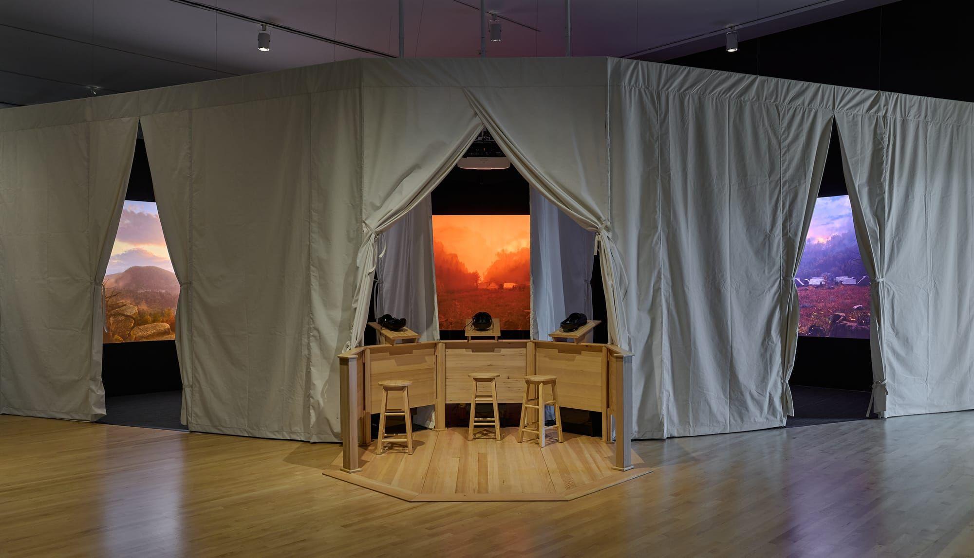 Panoramic view of a dimly lit installation featuring a large canvas tent with three openings. The polished wooden floor reflects the space. Each opening contains a screen displaying a moody, ominous landscape. In the center opening, a semicircular bar with three stools holds three VR headsets.