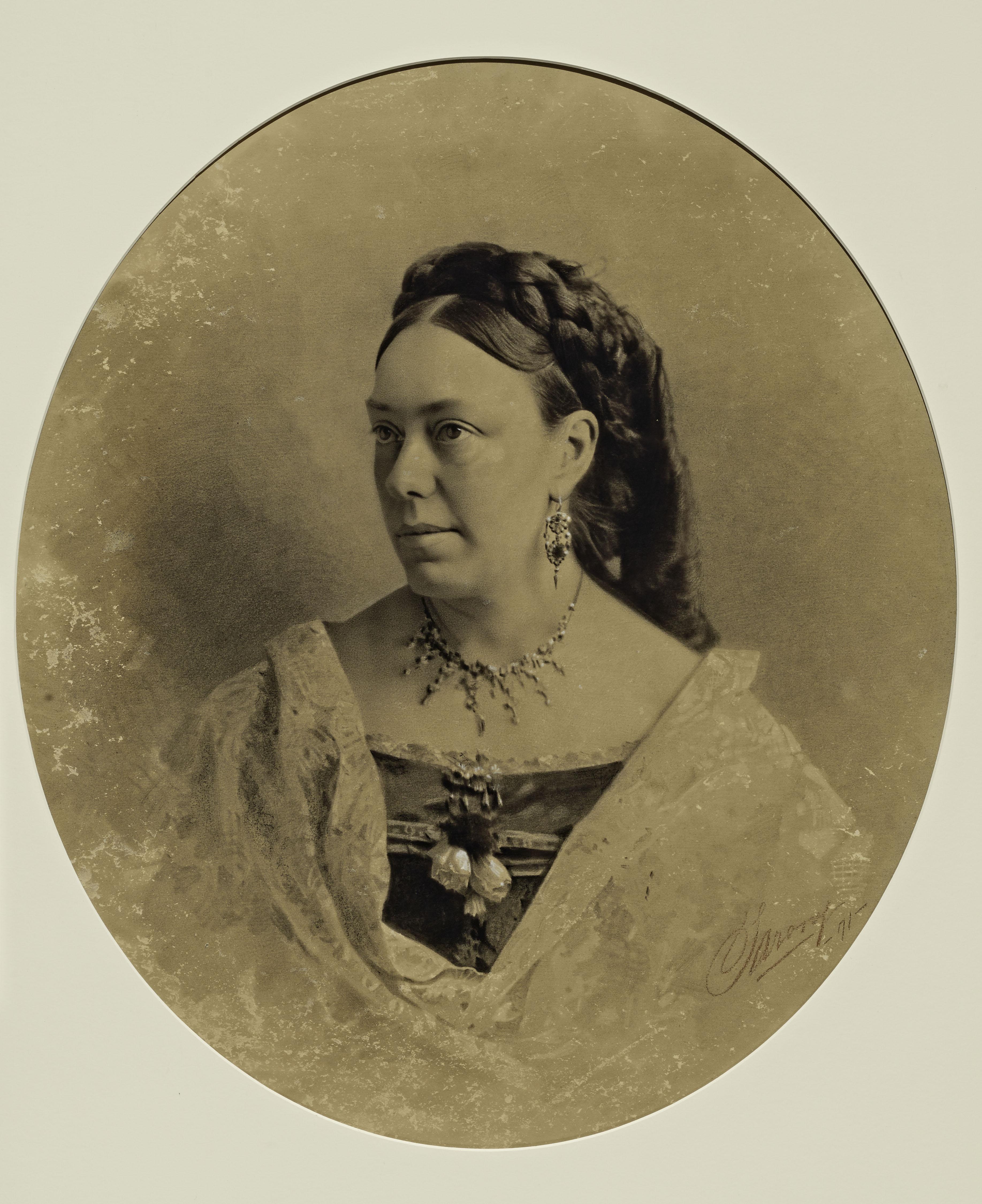 Short Alt Text A sepia-toned oval studio portrait of a woman looking to the side, wearing a formal off-the-shoulder dress, fine jewelry, and an elaborate braided hairstyle.
