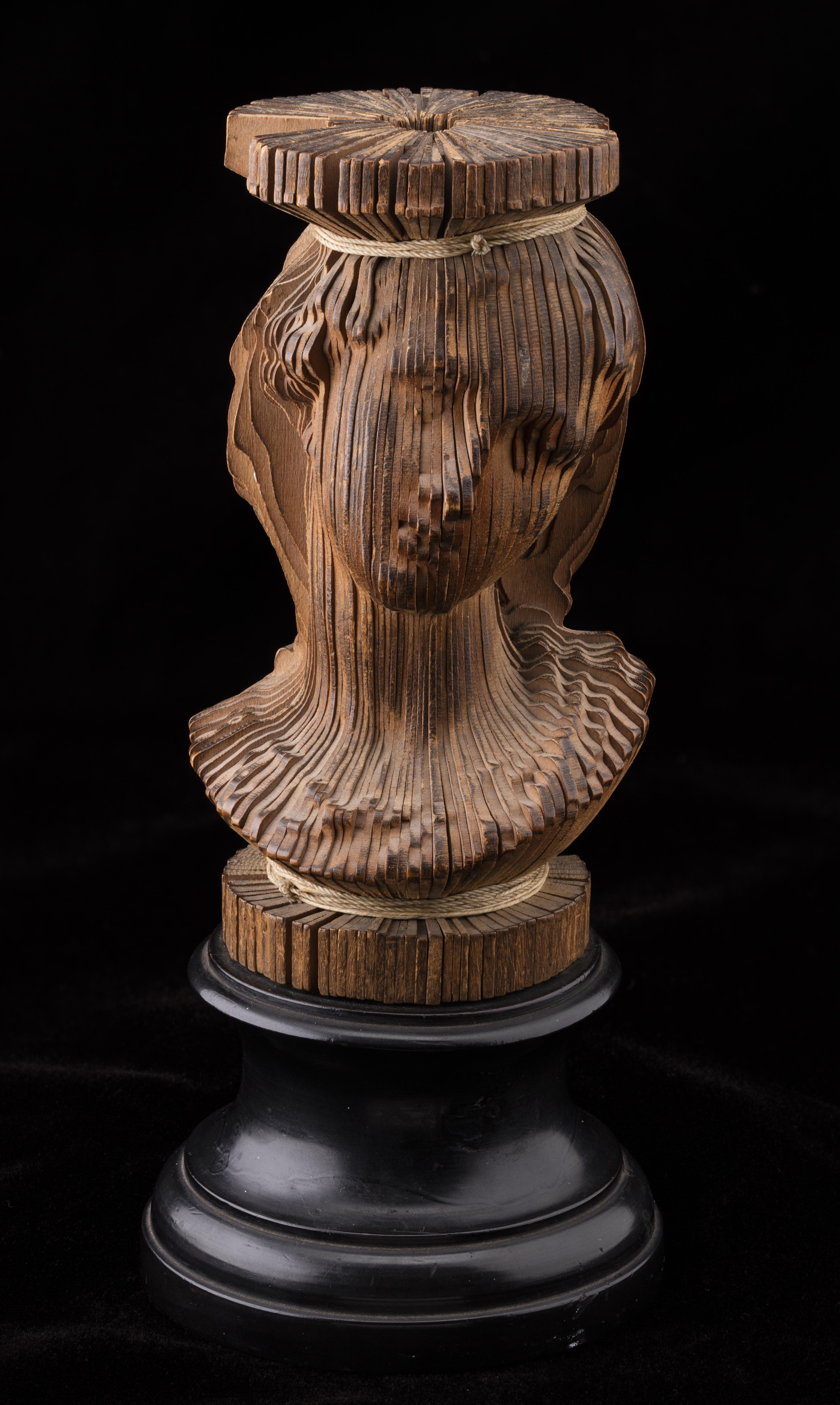 A wooden portrait bust sculpture set against a plain black background, mounted on a round black pedestal. The sculpture is constructed from numerous thin, vertical wooden slats arranged radially around a central axis, bound together near the top and base with thin light-colored cord. The carved edges of the slats collectively form the detailed contours of a person's head, face, and shoulder area.